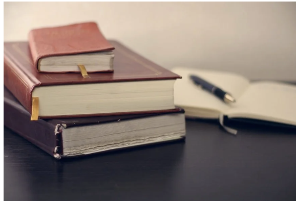 Stack of books beside an open notebook and pen on a desk.